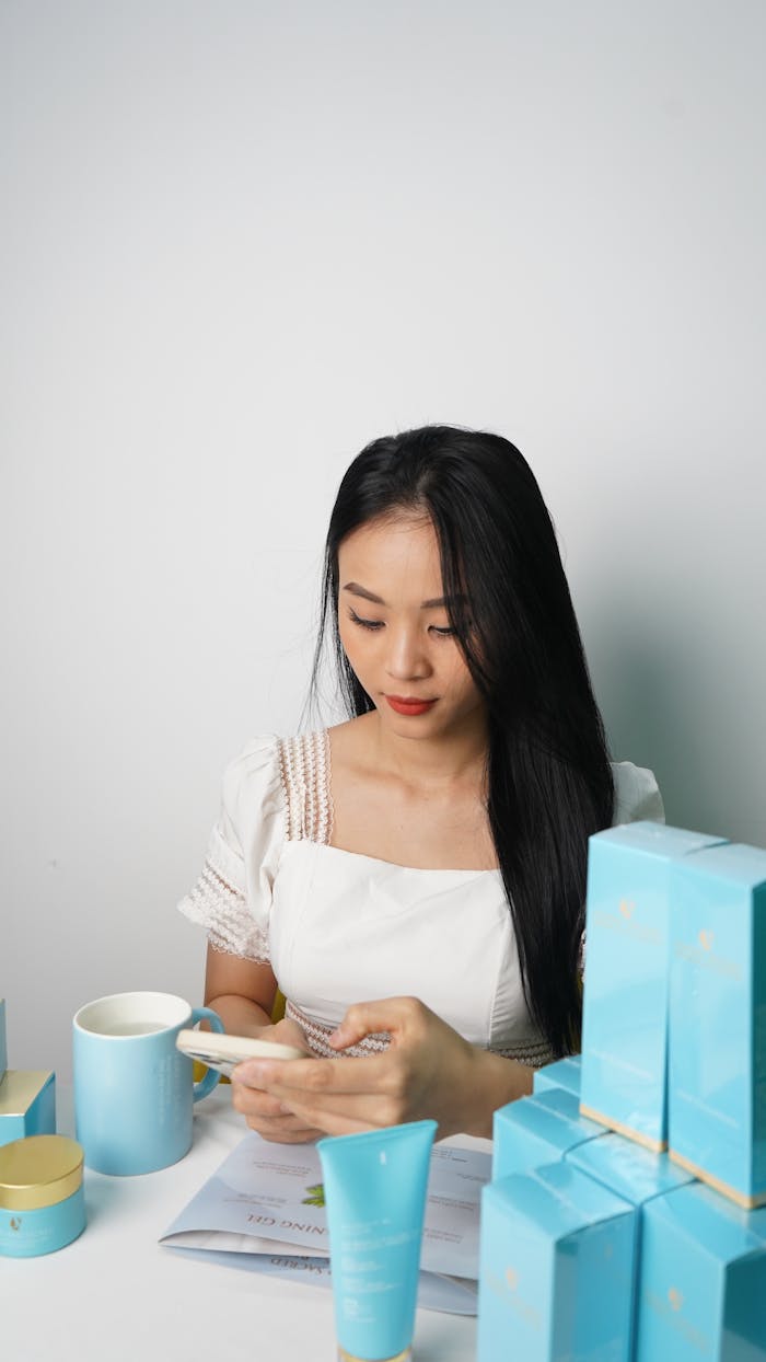 Young woman using smartphone surrounded by skincare products, showcasing modern beauty business.